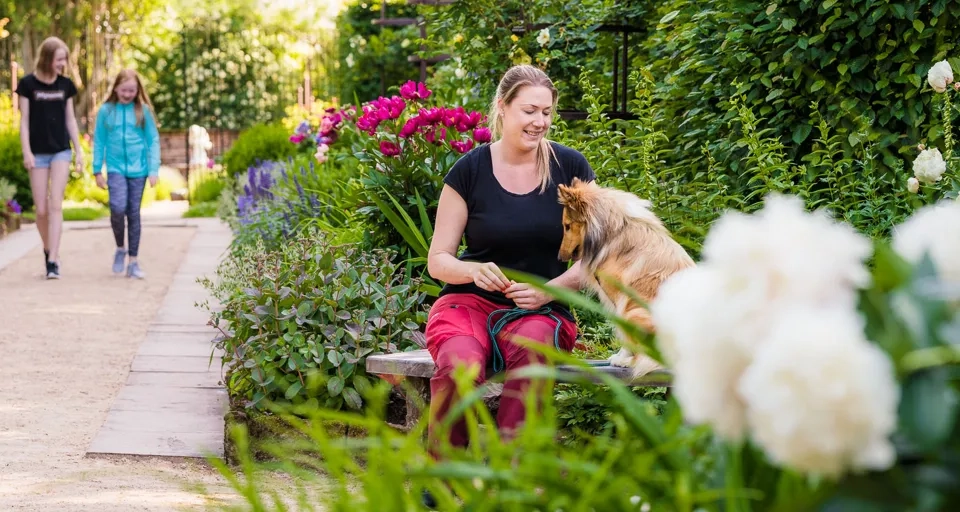 En slottsträdgård med blommor och en promenadgång omgiven av perenner. Två barn promenerar i bakgrunden och en kvinna sitter på en bänk och pratar med sin hund.