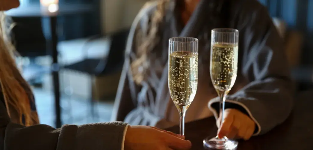 Två kvinnor står vid bardisk i badrockar med varsitt glas bubbel