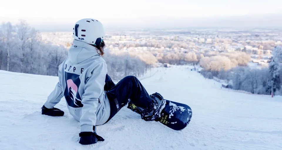 Person med hjälm och snowboard sitter i Billingebacken och tittar ut över backen och staden Skövde