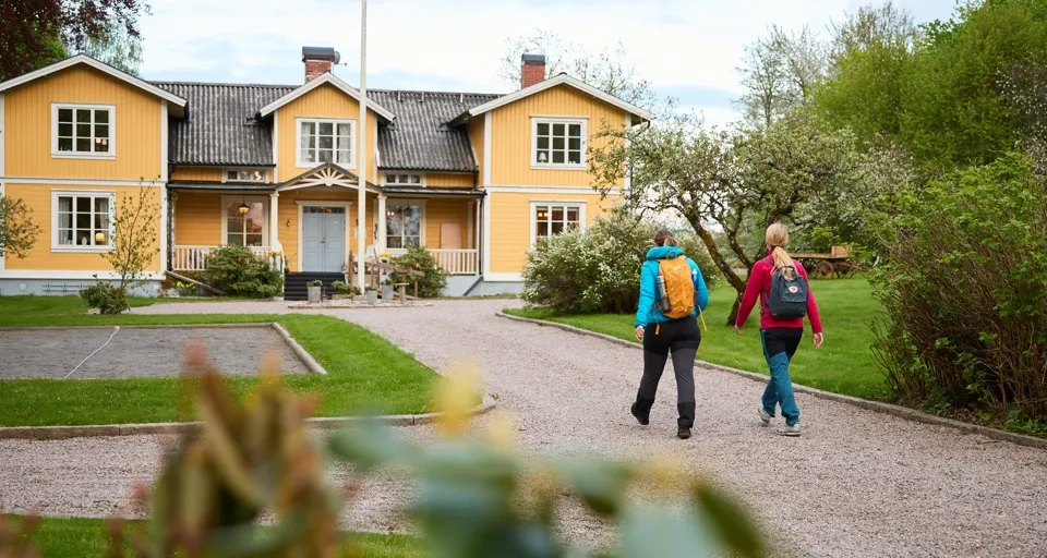 Två vandrare promenerar på uppfarten mot ett gult pensionat.