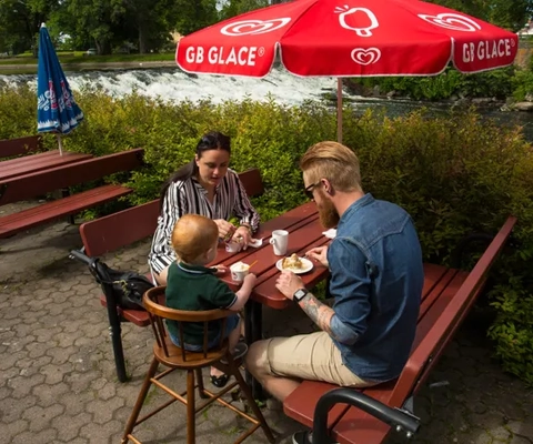 Två vuxna och ett barn sitter och fikar på terassen utmed ån Tidan på Turbinhusön i Tidaholm en sommardag.