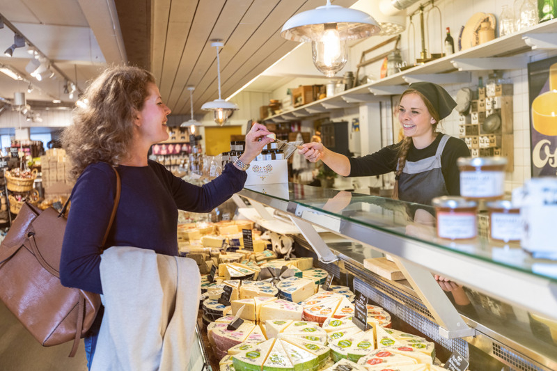 Kvinna som provsmakar en ostbit som expediten ger henne över en ostdisk.