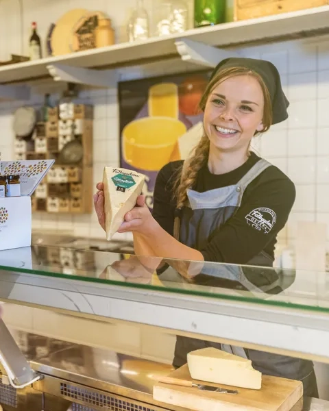 Kvinna som står bakom en ostdisk och håller upp en ost för en kund.