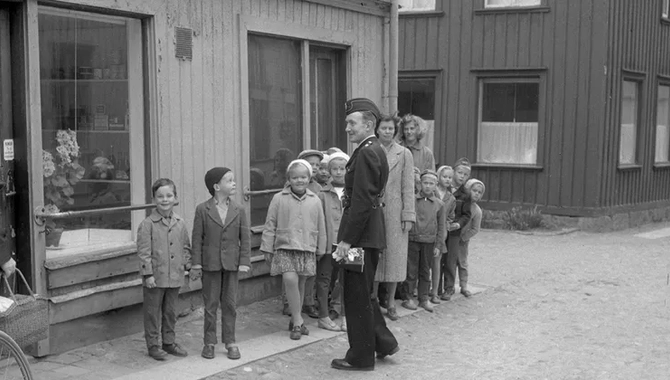 Flera barn och några vuxna står vid sidan av vägen intill en butik med en skylt om Charkuteri.