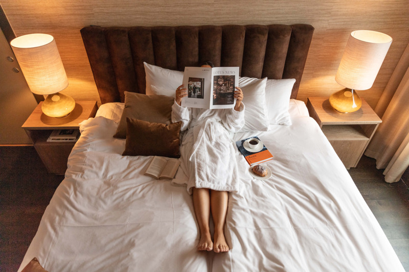 Person lying in a hotel bed reading a newspaper.