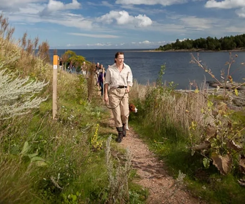 En kvinna i förgrunden och bakom sig har hon ett led med vandrare. Stigen går längs med vattnet.
