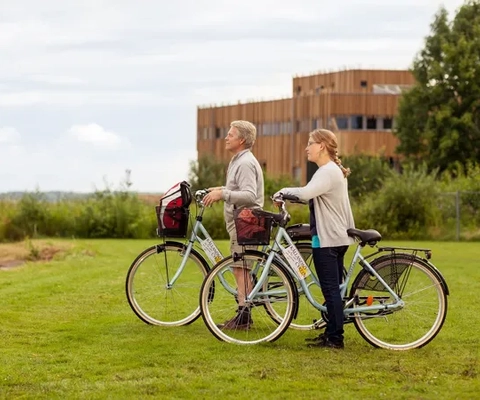 En man och en kvinna står med sina cyklar på en grön gräsmatta en mulig dag. I bakgrunden syns en brun träbyggnad.