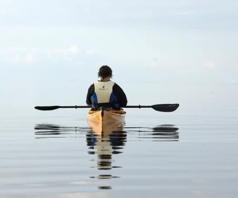 En person sitter i en kajak med ryggen mot kameran. Vattnet är stilla och går i samma gråblåa ton som himlen.