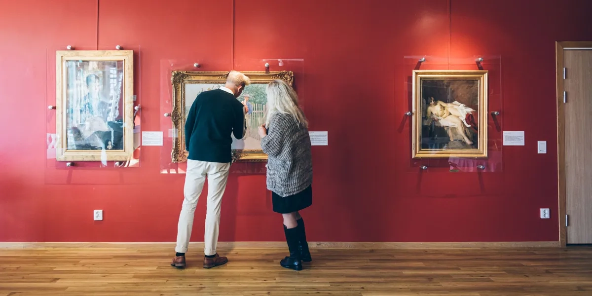 Två personer tittar på konst som hänger på en röd vägg.