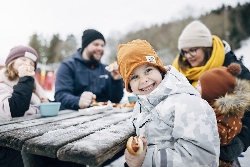 Familj sitter och äter korv vid ett bord utomhus. Pojke tittar rakt in i kameran och ler. 