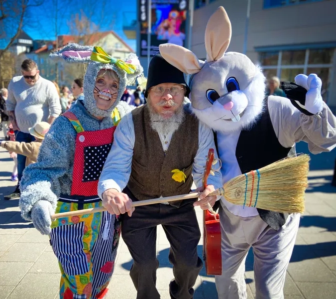 På rad står två påskharar med en påskgubbe i mellan sig. De håller en kvast framför sig. Bakom som syn ett folkvimmel.