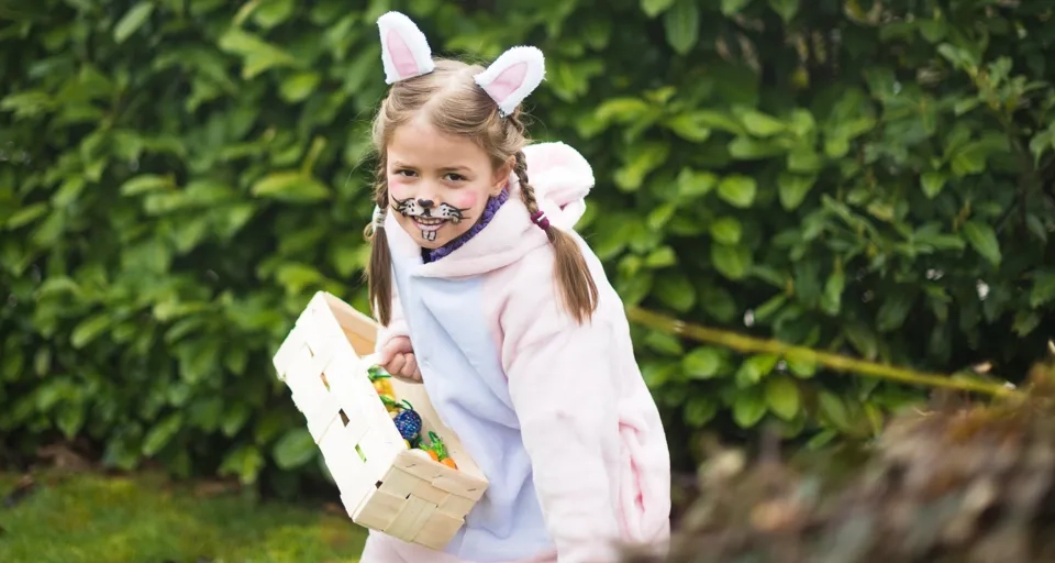 Ung flicka iklädd rosavit kanindräkt påväg mot kameran, håller i en korg med påskägg