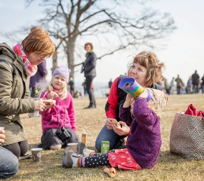 Barn och vuxna som sitter på marken och fikar. Ett av barnen kikar mot himlen med kikare.