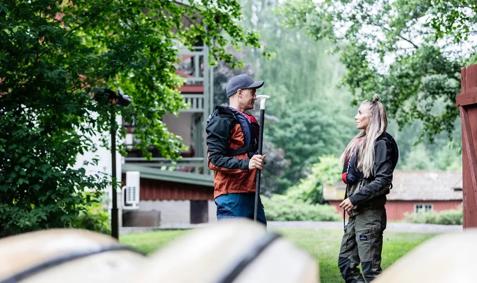 En kvinna och en man står i med var sin paddel i förgrunden syns några kanoter.