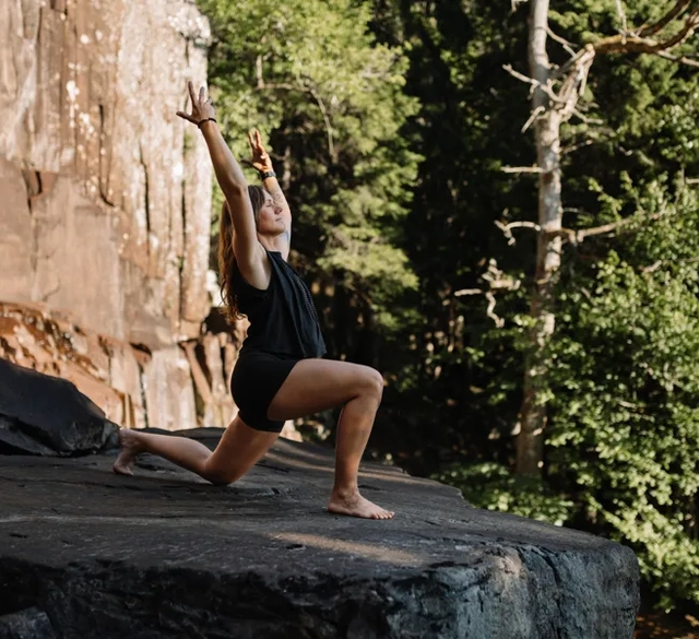 En kvinna yogar på en klippa intill en skog.