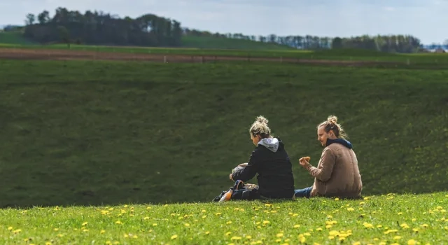 Två personer sitter på kanten till en dödisgrop täckt av grönt gräs. I förgrunden syns gula maskrosor.