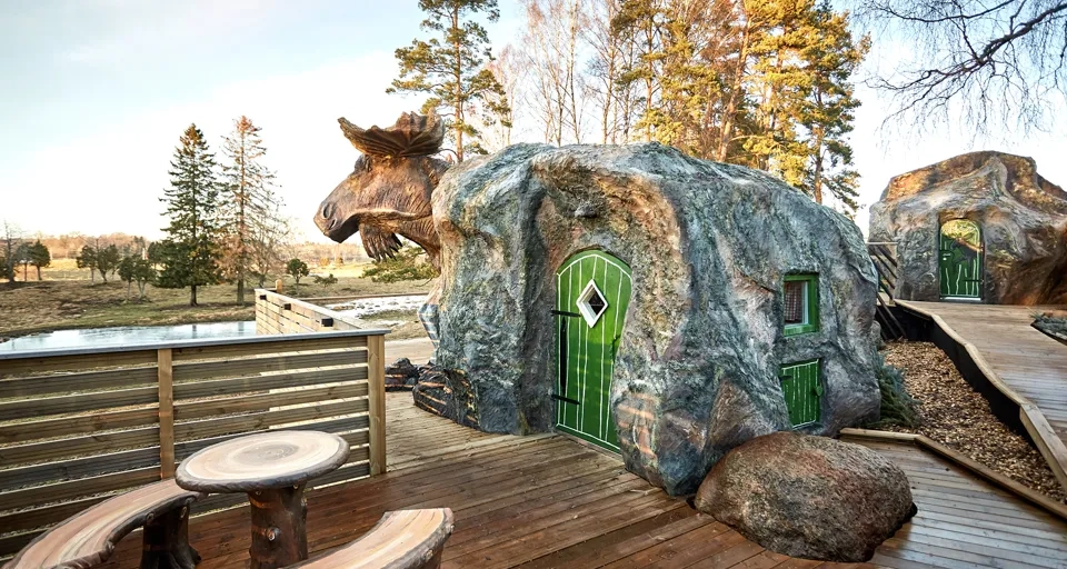 En stuga som ser ut som en älg. Gröna dörrar och bruna sittbänkar utanför.