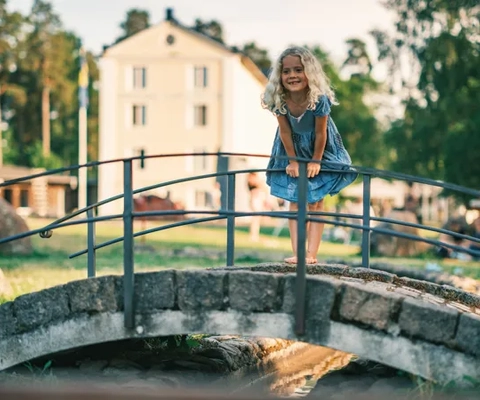 Flicka i blå klänning står vid räcket till en bro över minikanalen med Norrqvarns gula hotellbyggnad i bakgrunden.
