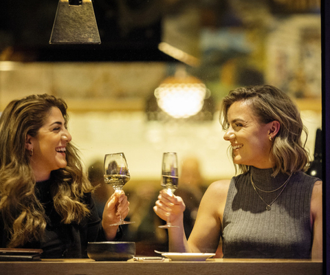 Två kvinnor sitter vid ett bord placerat vid ett fönster. De höjer varsitt glas mot varandra för att skåla.