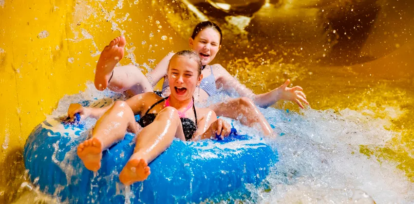 Två superglada tjejer åker på en blå badring i en gul vattenrutschkana så att vattnet stänker.
