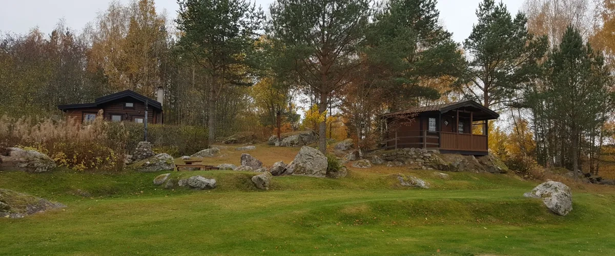 Vy över Camp Fjället utanför Tidaholm. Två bruna trästugor i skogskanten i höstskrud.
