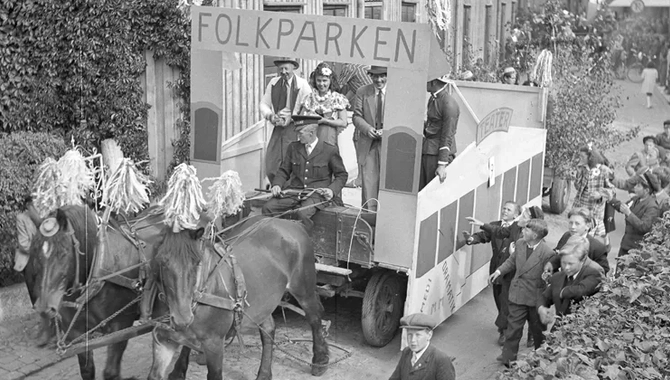 Två hästar med vagn och skylt om folkparken. Människor går i parad bakom vagnen. Byggnader i bakgrunden.