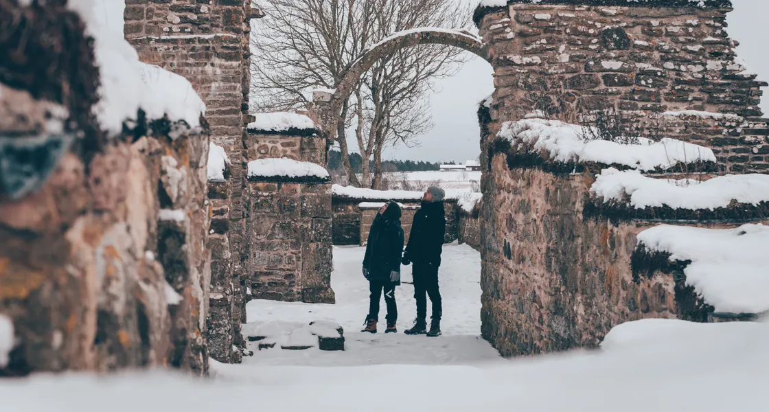 Snöigt landskap. Två personer står och kikar uppåt på en valvbåge i ruinerna av klosterkyrkan i Gudhem.