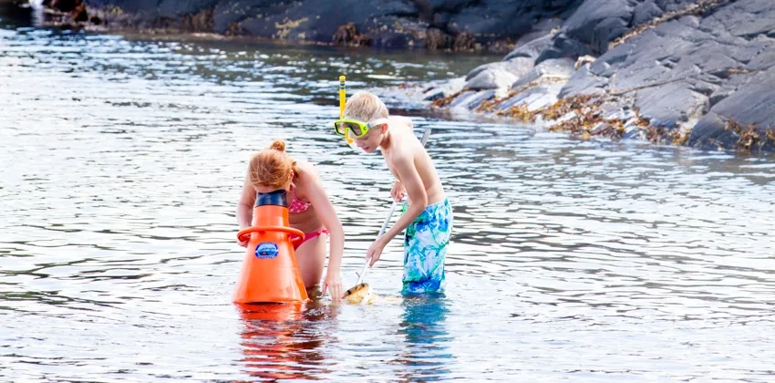 Två barn står i vattnet vid en strand. Ett av barnen har en stor, orange undervattenkikare och tittar ned på strandbottnen. Det andra barnet har en fiskehov.