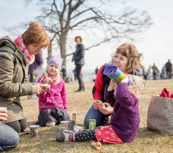 Barn och vuxna som sitter på marken och fikar. Ett av barnen kikar mot himlen med kikare.