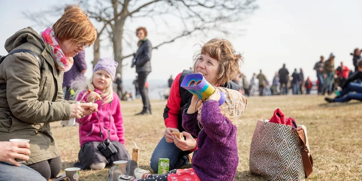 Barn och vuxna som sitter på marken och fikar. Ett av barnen kikar mot himlen med kikare.