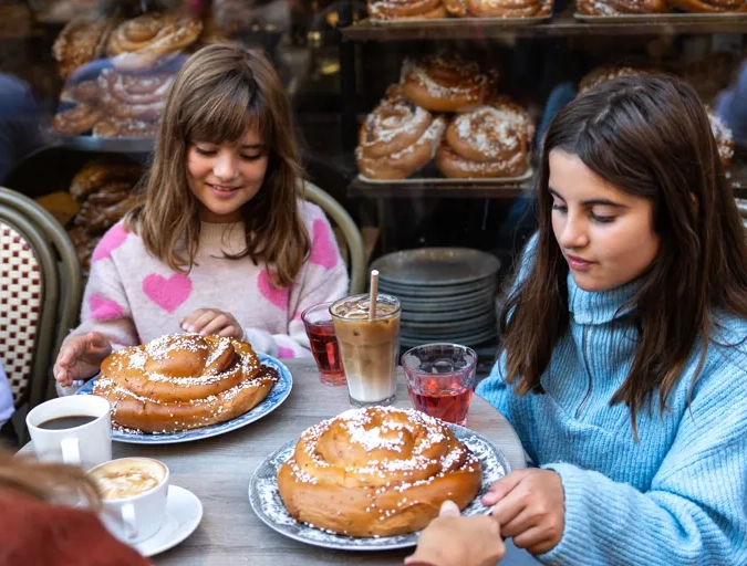 Två flickor som sitter runt ett bord, utomhus på ett kafé och dricker saft och äter kanelbullar