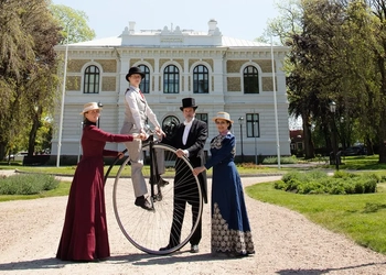 Fyra personer i 1800-tals kläder. En sitter på en höghjuling.