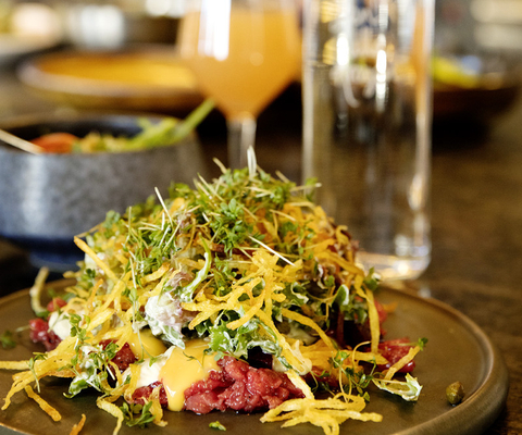 En tallrik med råbiff och tillbehör fint upplagt på råbiffen. I bakgrunden syns en drink, en vattenflaska och kocken.