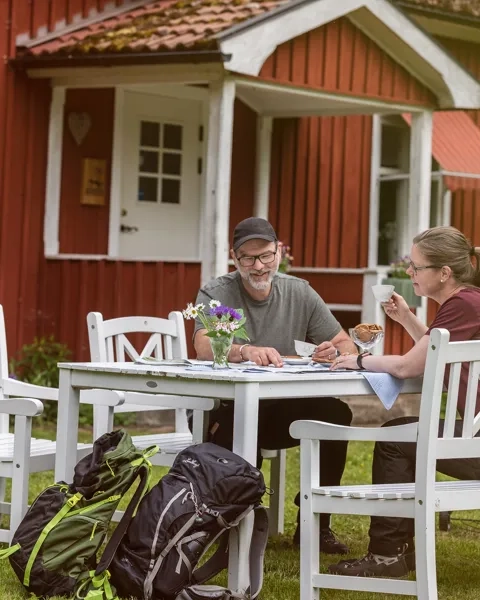 Röd stuga. Framför stugan sitter ett par och fikar i vita trädgårdsmöbler.