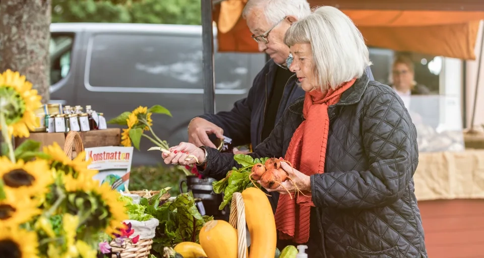 Man och kvinna som står och kikar på olika grönsaker på en marknad utomhus.