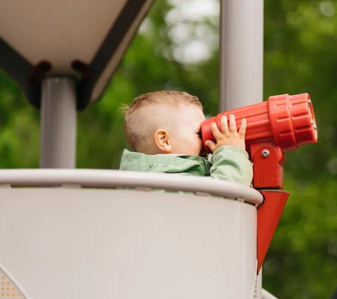 Pojke som står i en klätterställning och kikar i en röd plastkikare.