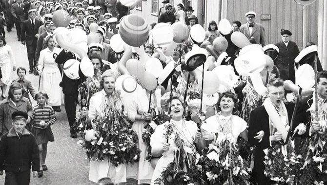 En parad med människor som firar student på en väg med hus i bakgrunden. De bär blommor runt halsen.