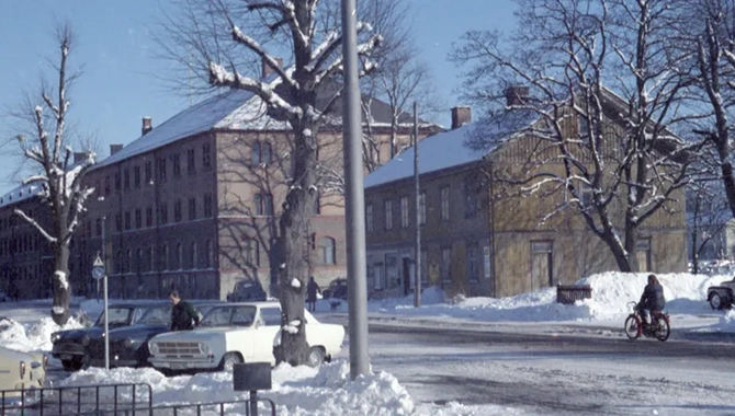 Snötäckt landskap. En väg med två parkerade bilar samt två mörka byggnader i bakgrunden. Träd står framför byggnaderna.