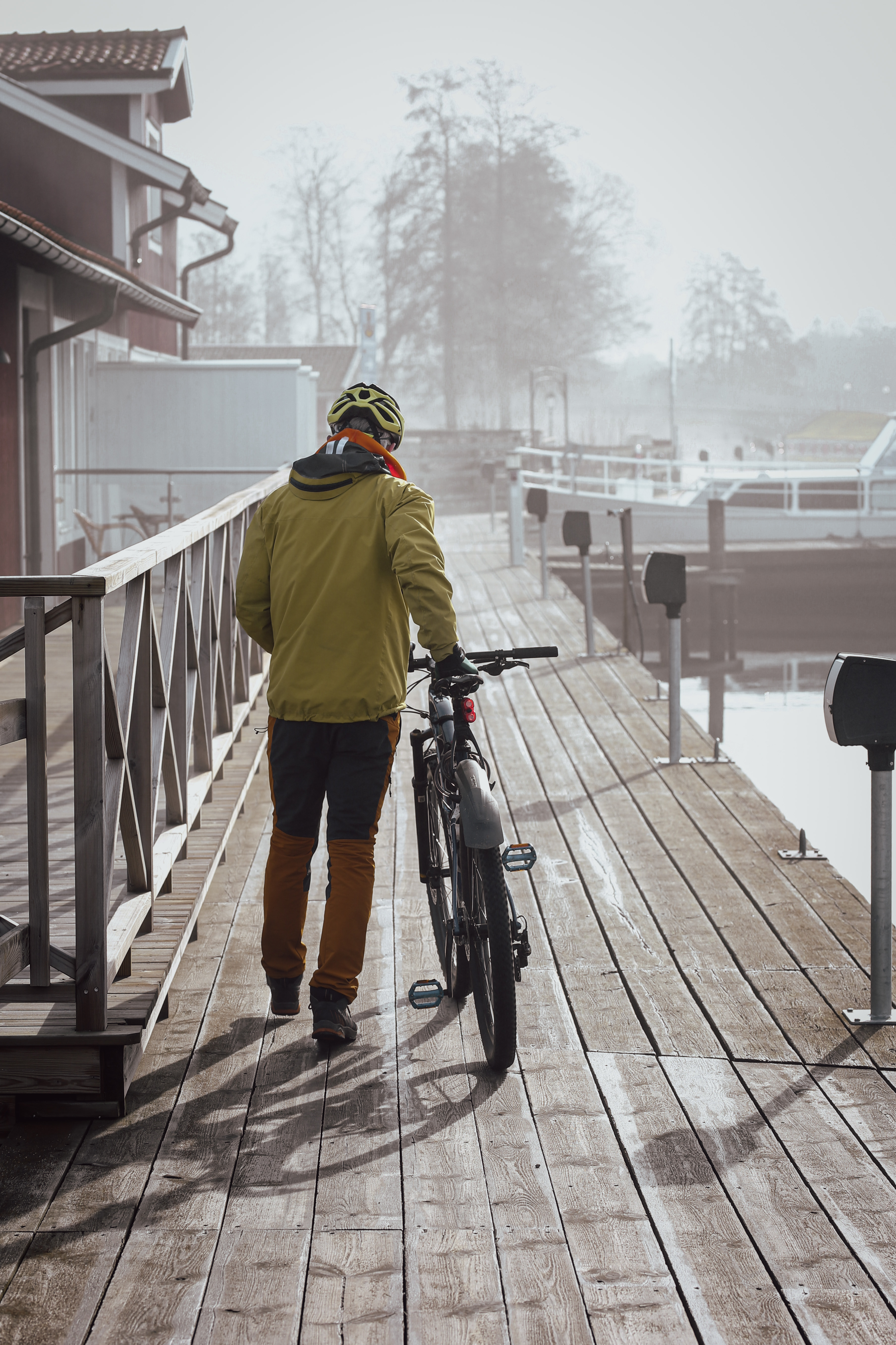 Man går från kameran ledande sin cykel. Han går på en brygga och i bakgrunden finns det träd. Sidan av ett hus syns på vänstersidan. 