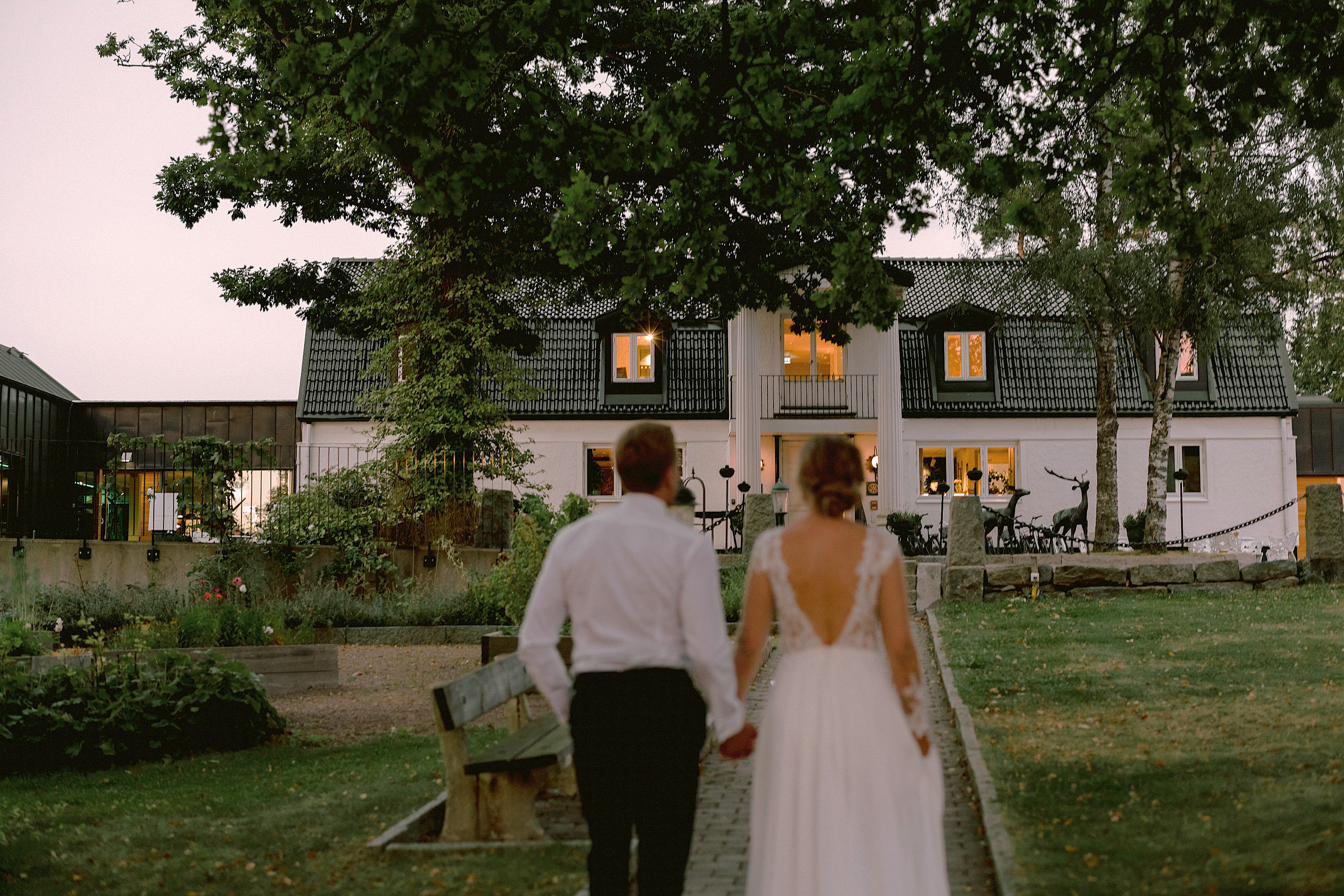 A bride and groom walk along a gravel path leading up to Hällsnäs Hotel