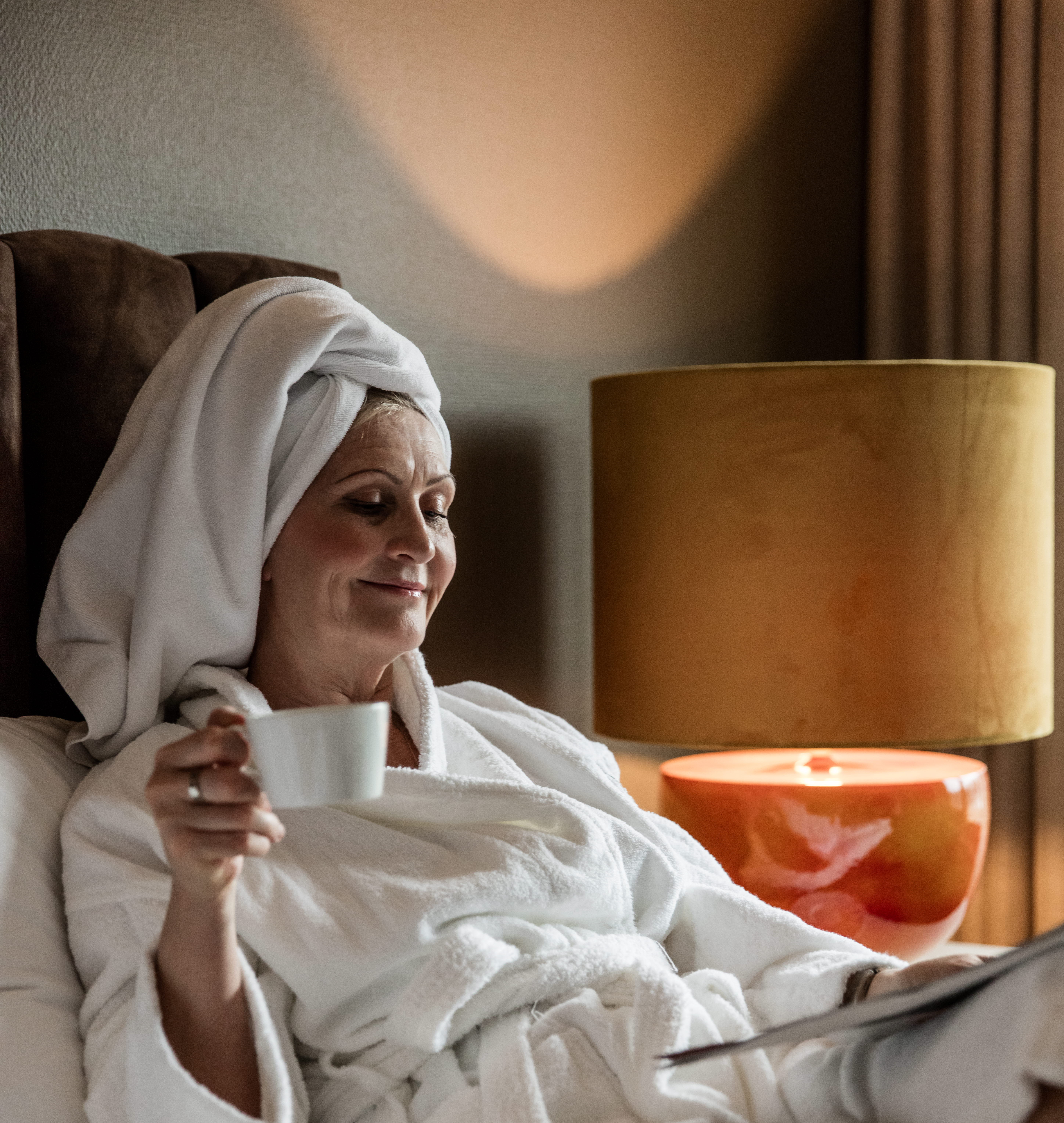 A hotel guest wrapped in a bath robe enjoying the comfortable bed and a cup of coffee.