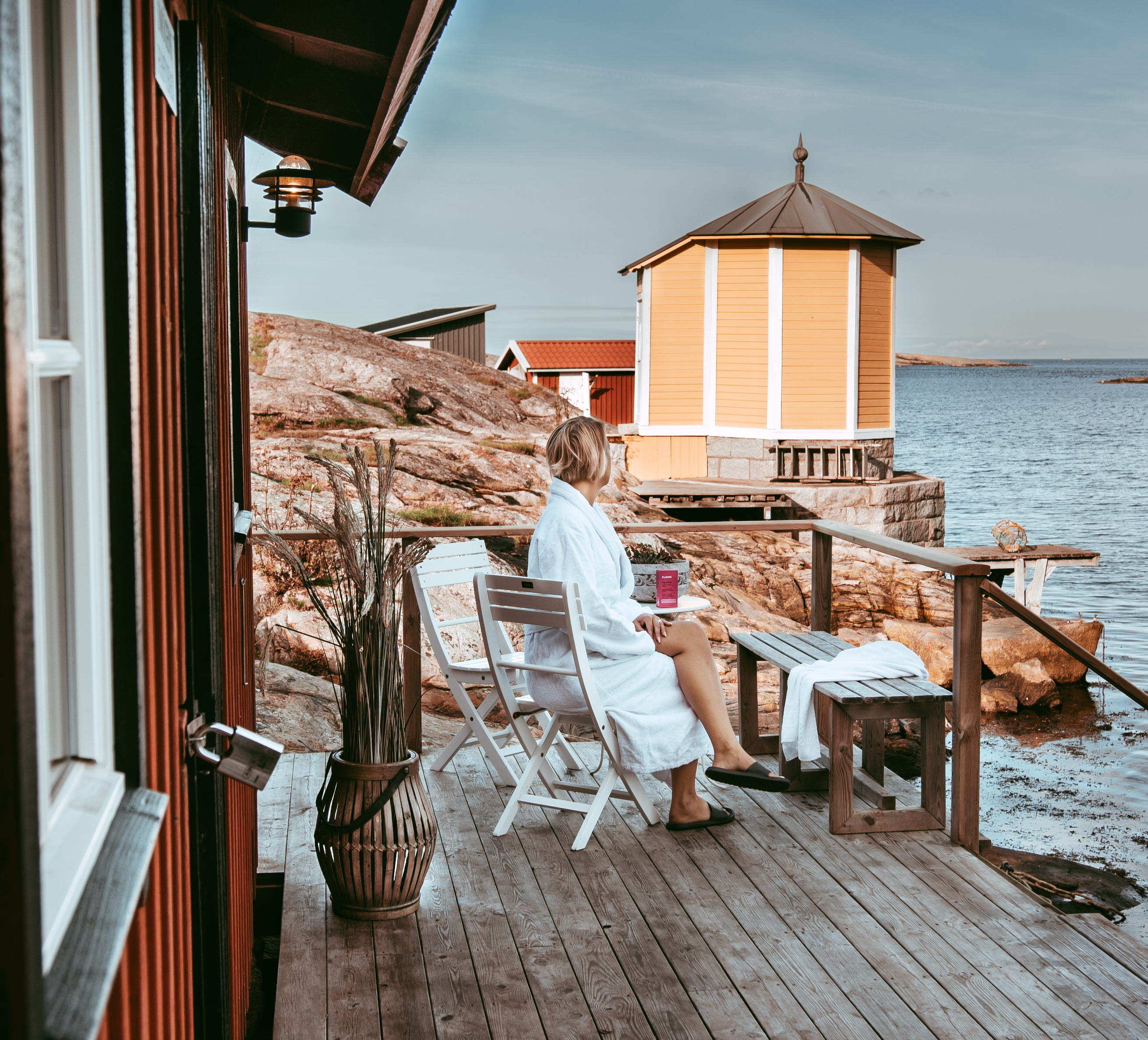 Woman in a bathrobe sitting outdoors at the bathing building in Makrillviken with the sea, bathhouse and rocks in the background, Hotell Smögens Hafvsbad