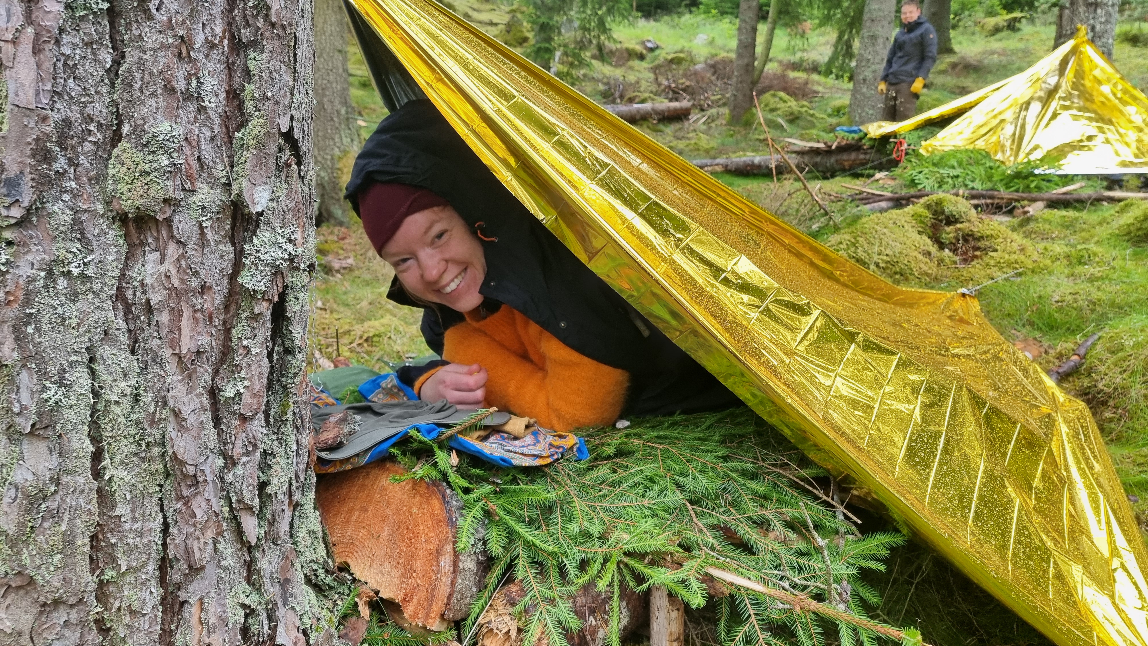 Överlevnadstält mot en tall och en leende kvinna på en bädd av granris