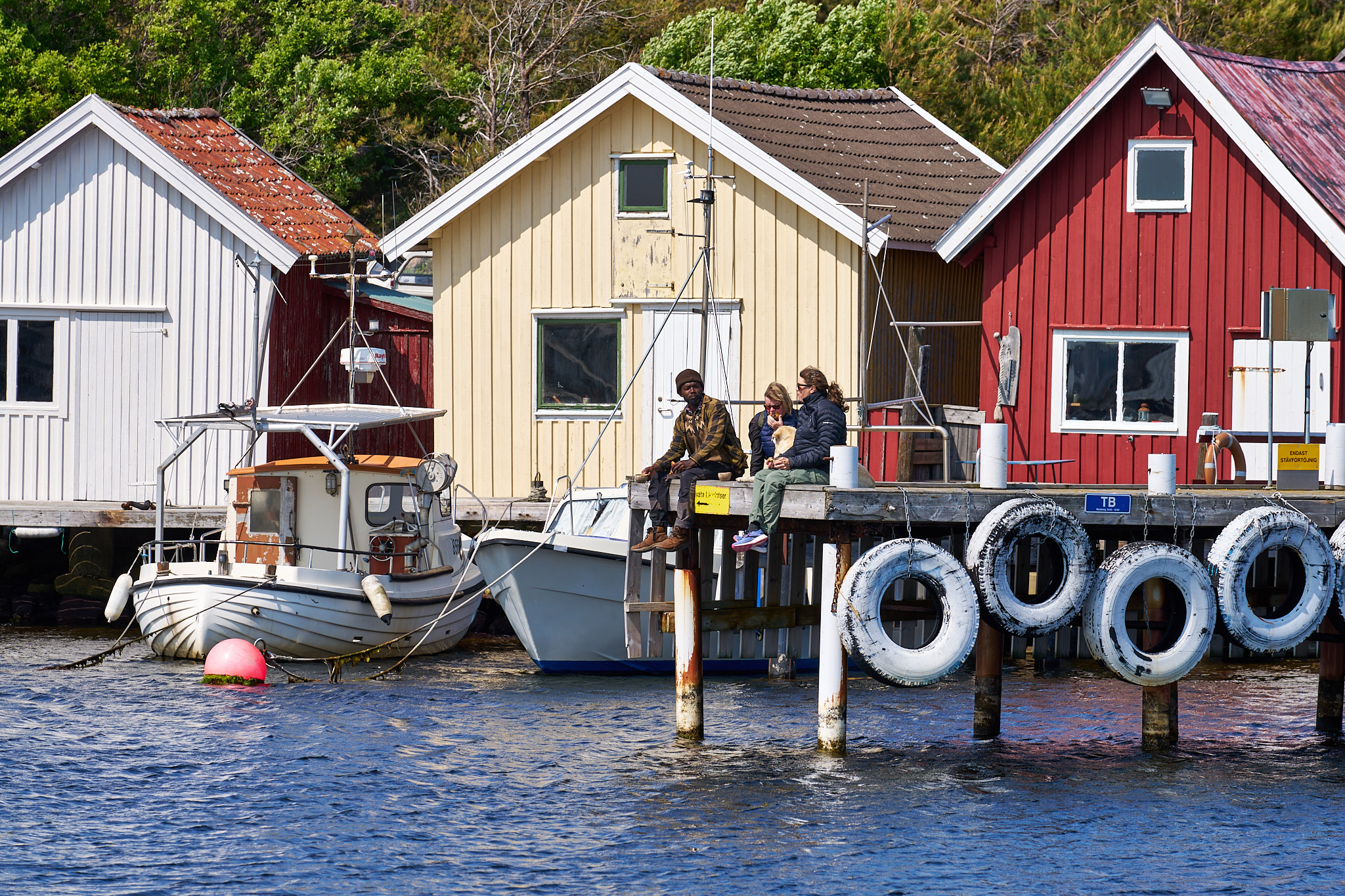 Three people have a coffee break on a dock in Brevik, in front of boathouses and a boat, in Kosterhavet National Par