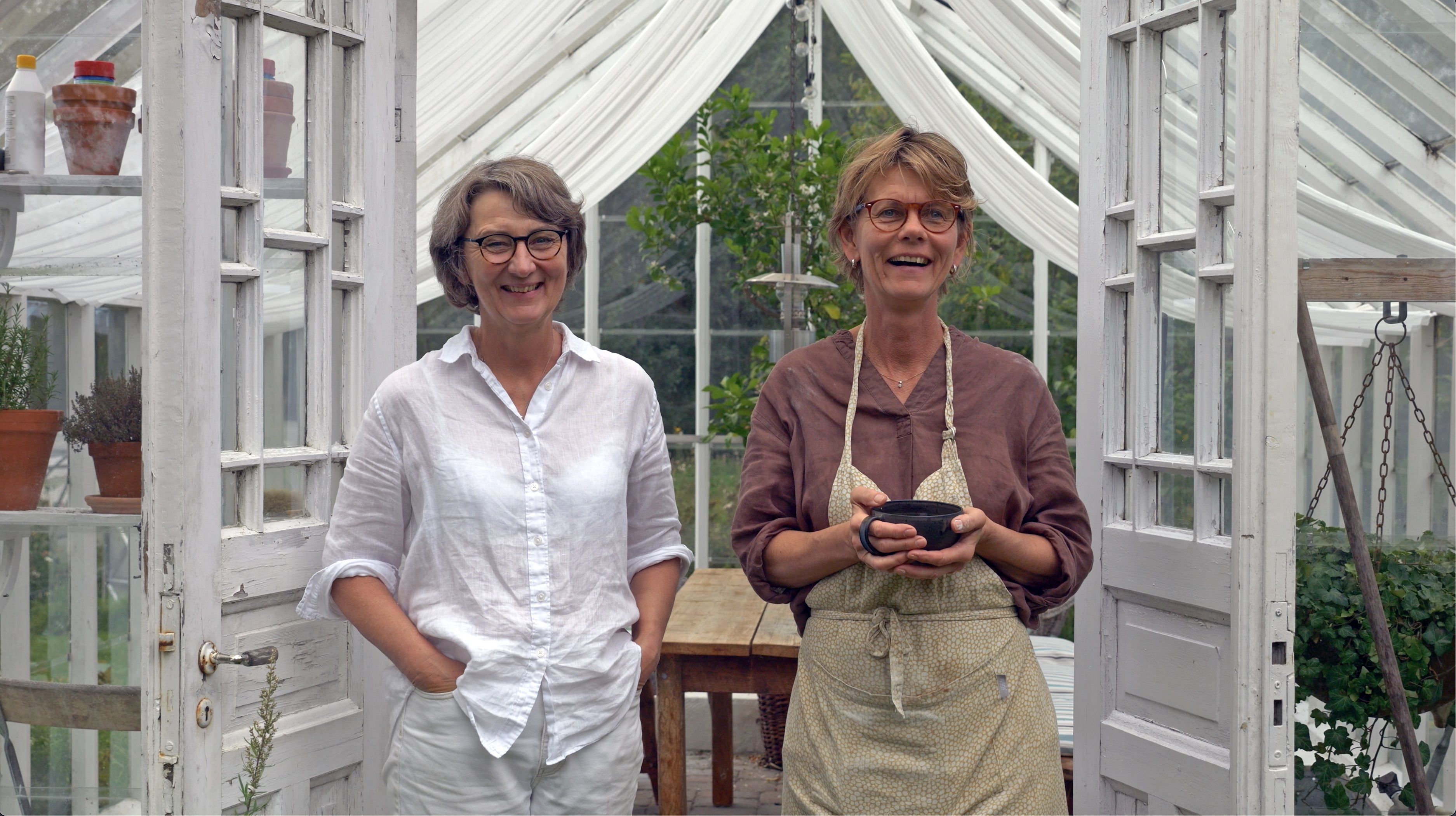 Two women stand in a greenhouse.