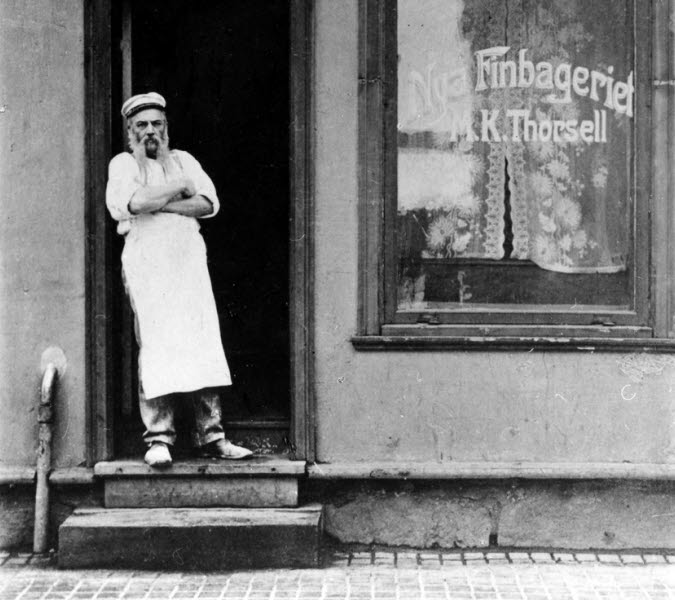 A baker stands in the front door of a bakery.