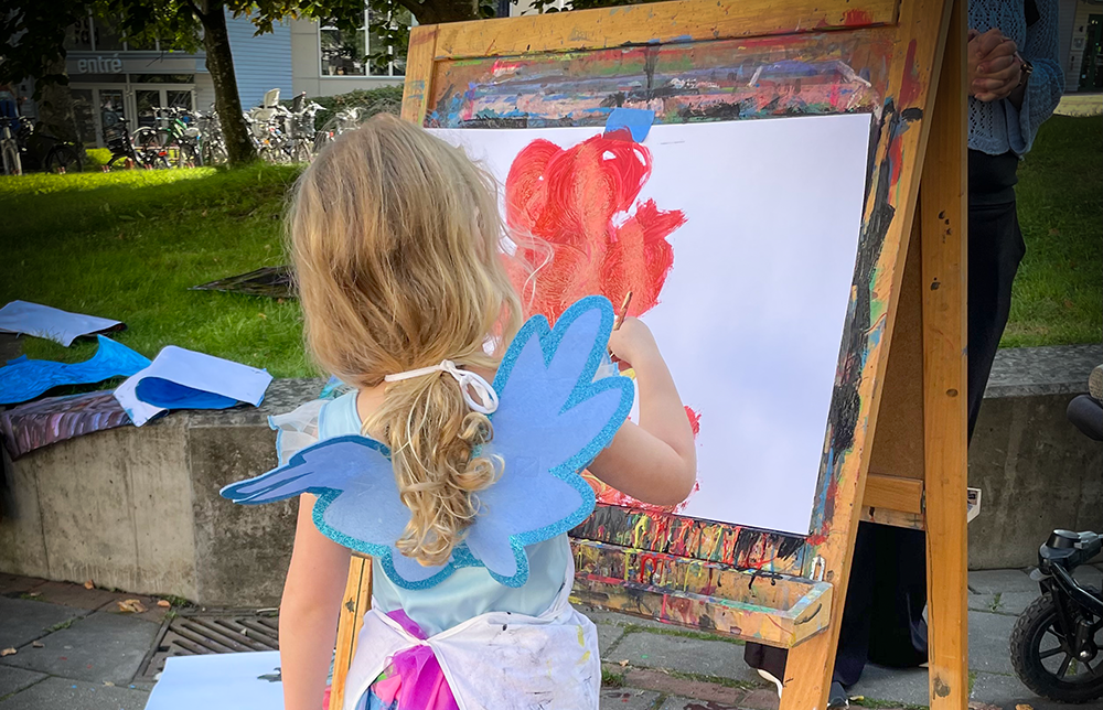 Barn med långt hår i en tofs målar med vattenfärg på staffli.