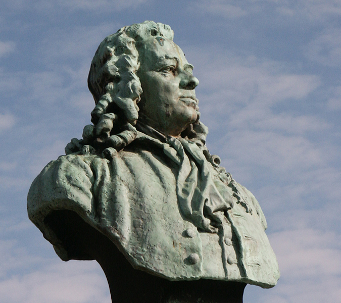 Staty av Jonas Alströmer mot blå himmel med slöjmoln. 