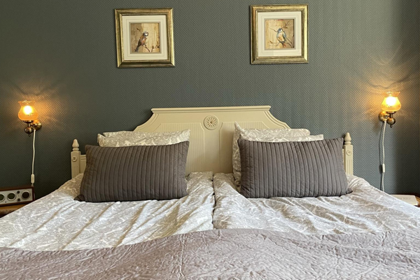 Dubbelsäng framför mossgrön målad vägg. I sängen ligger två bruna kuddar och ett beige överkast. På vardera sida om sängen finns två vägglampor. 