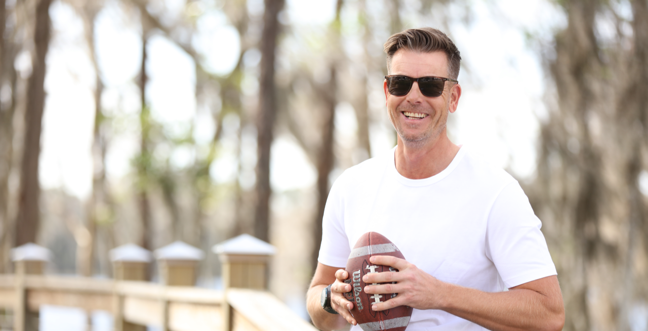 Man with a white t-shirt and sun glasses is sitting on a fence, holding an American football in his hands.